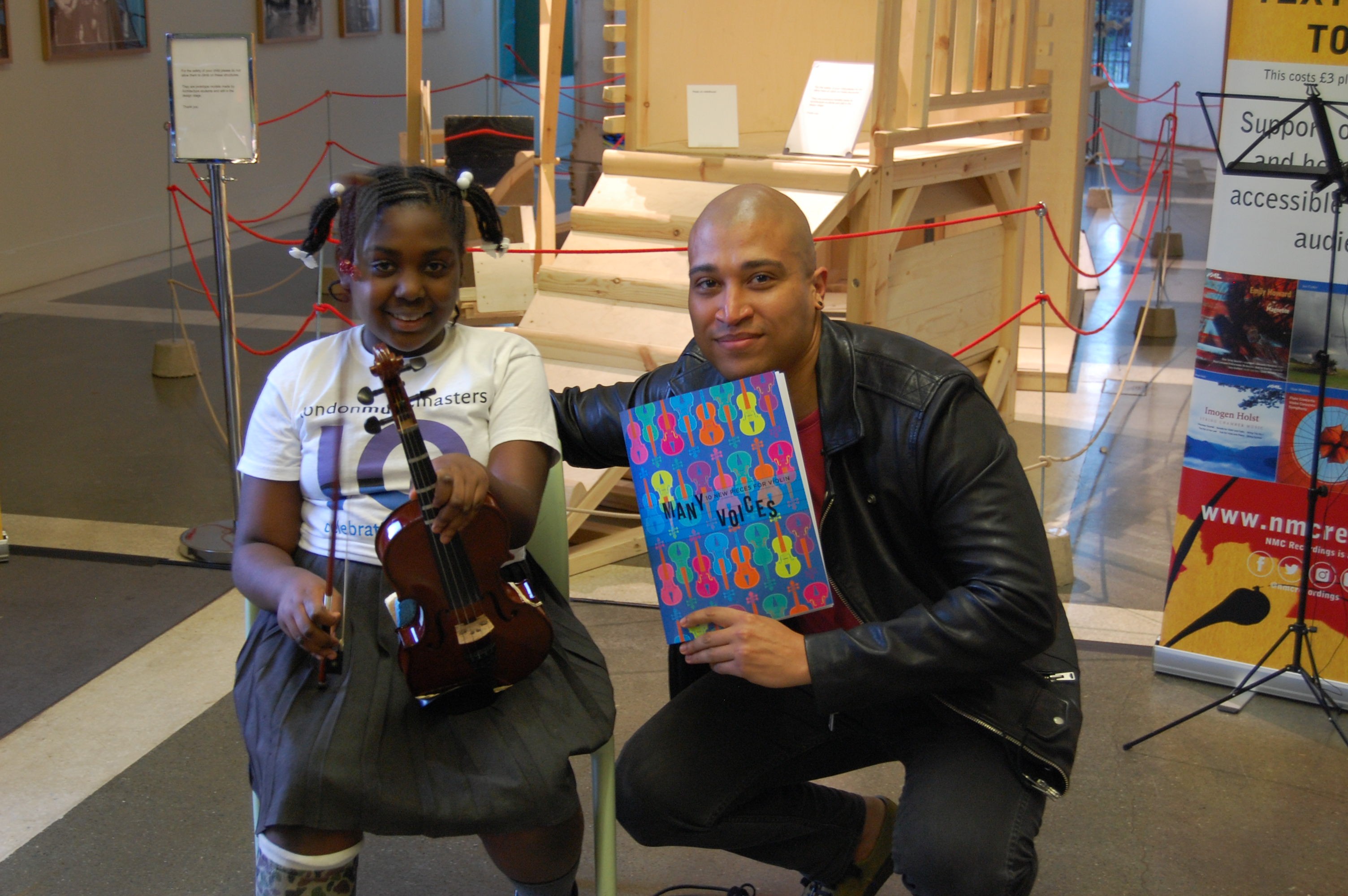 Composer Daniel Kidane holding the Many Voices book and posing for a photo with a young student holding a violin