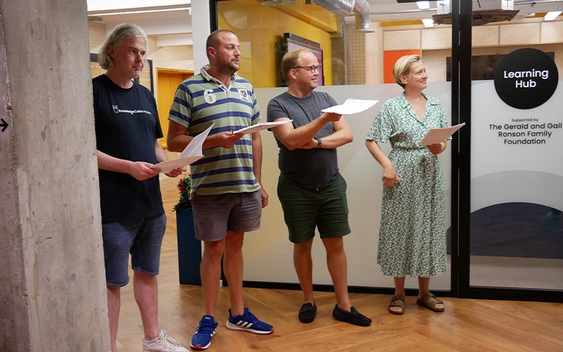 Four members of The Sixteen Choir stand together during rehearsal. They are holding scores, and are looking at someone off camera. They looked relaxed and happy.