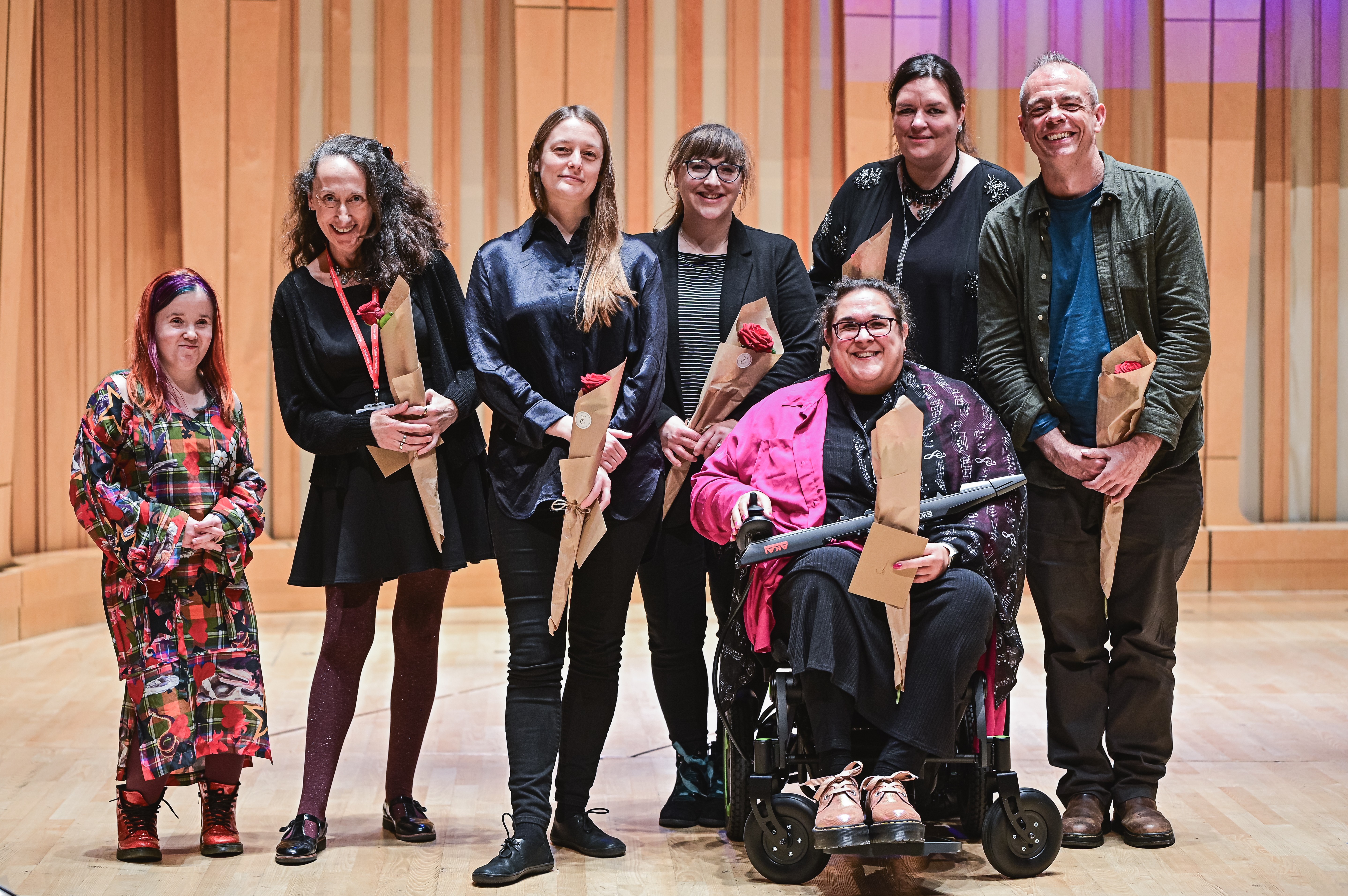 A group of seven composers, including one wheelchair user, stand on a stage holding wrapped bouquets. They are dressed in a variety of outfits, and the background is a studio featuring vertical wooden panels.
